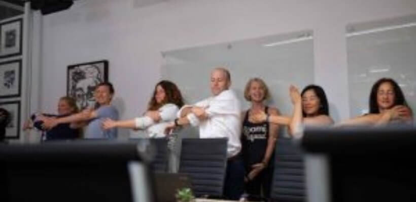 A group of diverse employees in an office are stretching their arms and shoulders. This image demonstrates an office wellness program, highlighting a focus on employee fitness, well-being, and healthy workplace habits.