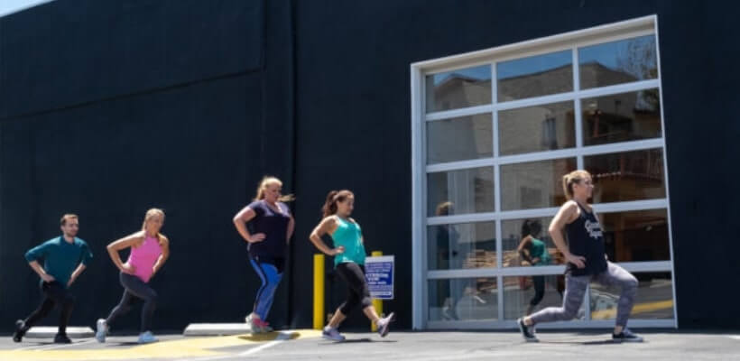 An outdoor group fitness class with five people doing lunges in a line. This image represents a corporate wellness program, employee health initiative, and workplace exercise.